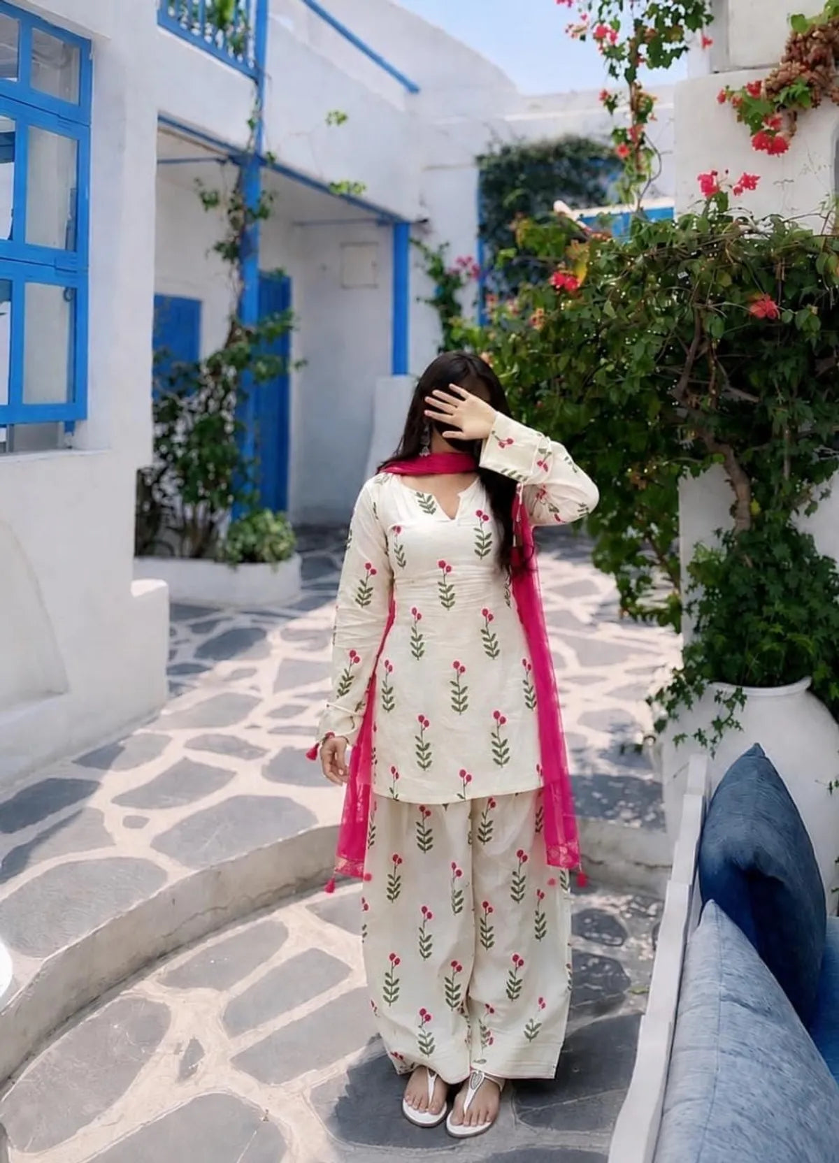 A woman wearing a cream-colored embroidered salwar kameez suit with a bright pink dupatta, standing in a scenic outdoor setting with white buildings, blue accents, and green foliage.