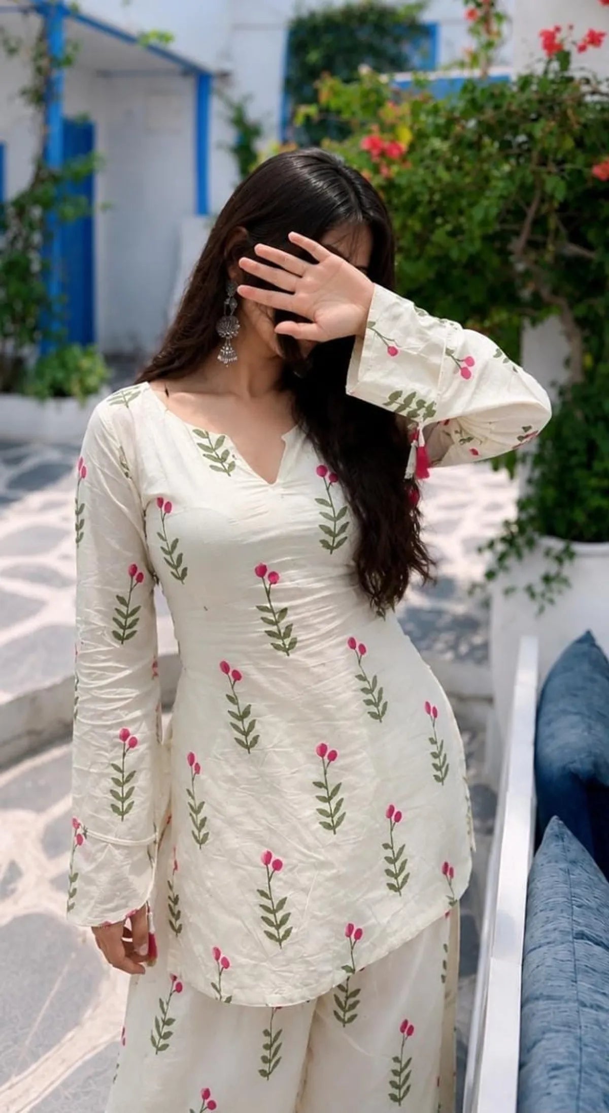 A woman wearing a cream-colored kurta set with pink floral prints and green leafy stems, posing outdoors in front of a white wall with blue accents and green plants.