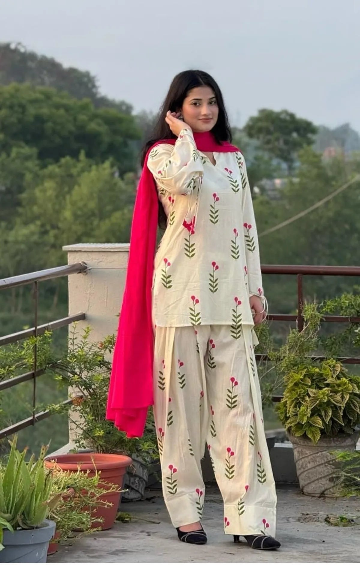 A woman stands on an outdoor balcony wearing a cream-colored embroidered kurta and pant set with a bright pink dupatta. The outfit features green leaf patterns and small pink floral accents.